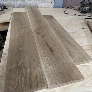 May include: Three unfinished wooden planks with a light brown grain pattern. The planks are rectangular and appear freshly cut, ready for a woodworking project. The wood has a natural, rustic appearance.