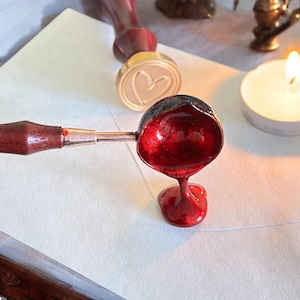 May include: A red wax seal being poured from a spoon onto a white envelope. A wooden handle is attached to the spoon. A gold stamp with a heart design is in the background.