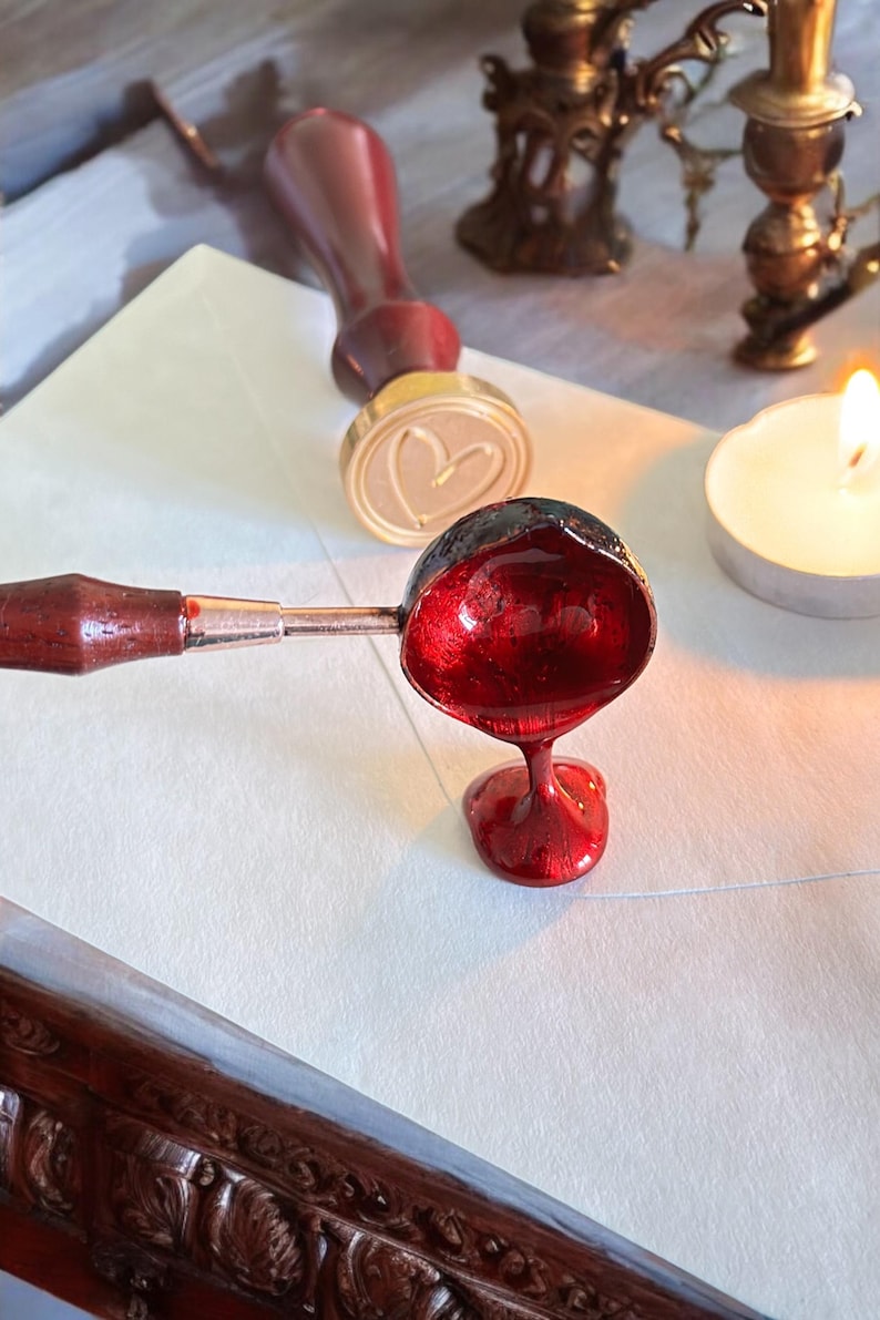 May include: A red wax seal being poured from a spoon onto a white envelope. A wooden handle is attached to the spoon. A gold stamp with a heart design is in the background.