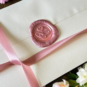 May include: A cream envelope sealed with a pink wax seal, featuring a heart and ring design. A pink ribbon is tied around the envelope, and the word "SMYTHSON" is embossed. Flowers in the background add a decorative touch.