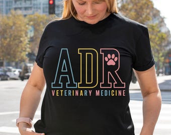 May include: Black t-shirt with the letters "ADR" in blue, yellow, and pink, with a paw print in the "R". The words "VETERINARY MEDICINE" are below the letters. The shirt is worn by a person.