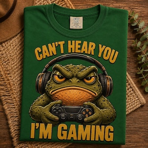 May include: A dark green t-shirt featuring a graphic of a textured, grumpy-looking toad wearing black over-ear headphones and holding a black game controller. The yellow text above reads CAN'T HEAR YOU and the text below reads I'M GAMING.