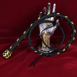 Könnte beinhalten: Eine schwarze und goldene geflochtene Lederpeitsche mit einem dekorativen Knoten am Ende. Die Peitsche ist um eine silberne Roboterhand gewickelt.