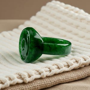 May include: A solid, deep green ring featuring a smooth, polished finish and a circular, slightly raised center design. The band is thick and uniform, showcasing natural variations in the green stone material throughout its entire structure.
