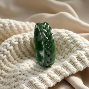 May include: A dark green jade ring with a braided design. The ring is set against a cream-colored knitted sweater and a beige fabric background. The ring's intricate braid pattern is clearly visible.