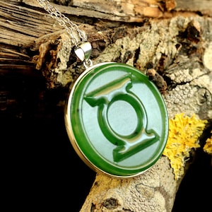 May include: A circular green pendant featuring the embossed Green Lantern symbol in the center. The pendant is encased in a silver-toned metal frame and hangs from a delicate silver-toned chain, showcasing a smooth, polished surface finish.