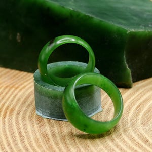Könnte beinhalten: Zwei polierte Jade-Ringe in leuchtendem Grün liegen auf einem kleinen grünen Ständer, mit einem großen Jadeblock im Hintergrund. Die Ringe sind glatt und kreisförmig und zeigen die natürliche Farbe und Textur des Jades. Die Ringe werden auf einer Holzoberfläche präsentiert.