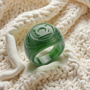 May include: A thick, polished dark green ring featuring a circular emblem with the Green Lantern logo embossed on the top surface. The band has a smooth, rounded profile with subtle carved details surrounding the central circular symbol.