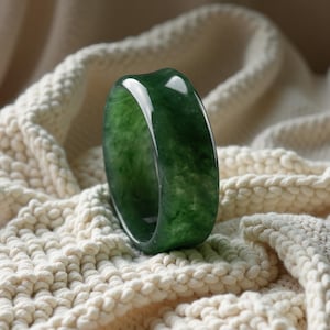 May include: A smooth, polished, dark green jade ring with a wide, rounded band. The stone features natural mottled patterns and variations in color intensity throughout its surface, reflecting light with a glossy, reflective finish.