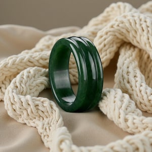 Könnte beinhalten: Ein dunkelgrüner Jade-Ring mit glatter, polierter Oberfläche. Der Ring hat ein gerilltes Design und ist vor dem Hintergrund eines cremefarbenen, gestrickten Textils platziert.