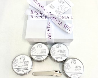 May include: A white gift box with a purple ribbon tied around it. The box contains four small silver tins with labels that say "Aroma Spa" and list different facial products. The products are: Facial Cleansing Balm, Facial Scrub, Warm Oil Face Mask, and Facial Cream. All products are labeled "Sensitive" and "100% Natural".