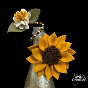 May include: A felt sunflower with a brown center is attached to a clear glass bottle with a beaded necklace. A smaller felt flower with white petals and yellow center is attached to the bottle.