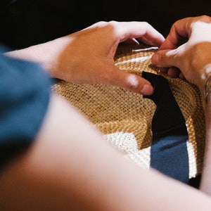 May include: A person is sewing a black ribbon to a straw hat. The hat is light brown and has a woven texture.