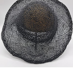 Könnte beinhalten: Ein schwarzer Sonnenhut mit breiter Krempe. Der Hut hat eine runde Krone und eine breite Krempe in offener Webart. Der Hut besteht aus einem dunklen Material, wahrscheinlich Stroh oder einer ähnlichen Naturfaser. Der Hut soll vor der Sonne schützen.