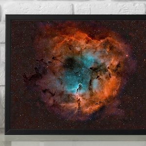Op de afbeelding: Een ingelijste foto van een nevel in de ruimte. De nevel is een wervelende wolk van gas en stof, met tinten rood, oranje en blauw. De nevel wordt omringd door een veld van sterren.