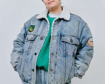 May include: A light blue denim jacket with a sherpa collar and several patches. The jacket has a green sweater underneath.