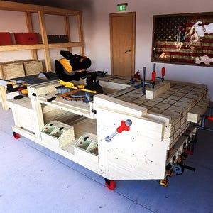 May include: A wooden workbench with a built-in miter saw, various tools, and red clamps. The workbench is on wheels and has storage shelves. An American flag hangs on the wall.