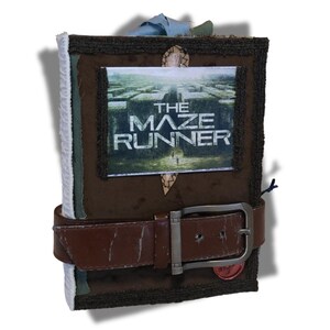 May include: A handcrafted journal with a brown leather belt fastening. The cover displays a framed image from "The Maze Runner" film. The journal has a textured, dark brown cover with decorative stitching and a fabric spine.