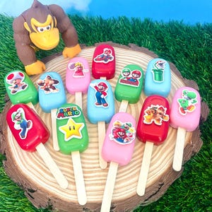 May include: A collection of colorful, character-themed popsicles on wooden sticks, featuring designs from the Super Mario franchise. The popsicles are arranged on a wooden slice, with a Donkey Kong figurine in the background. The popsicles are various colors, including red, green, blue, and pink.