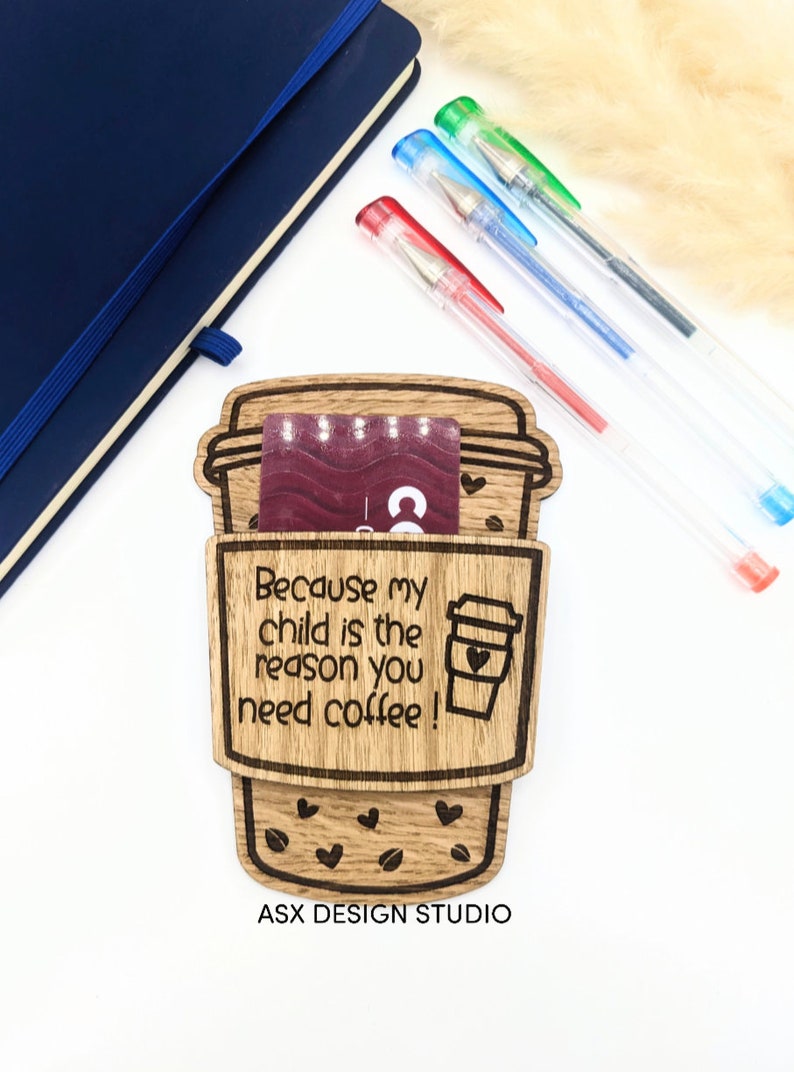 May include: Wooden coffee cup holder with the text "Because my child is the reason you need coffee!" and a small coffee cup with a heart on it. The holder has a slot for a gift card and is decorated with coffee beans and hearts. The text "ASX DESIGN STUDIO" is at the bottom.