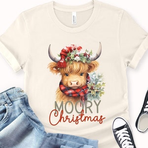 May include: A cream colored t-shirt with a cartoon illustration of a Highland cow wearing a red and black plaid scarf and a red Santa hat with holly. The text "MOORY Christmas" is printed below the cow.