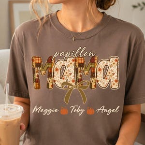 May include: A brown short-sleeved t-shirt featuring the text "papillon MAMA" in a patchwork-style font with autumn-themed patterns. A tan bow graphic sits below the text, followed by the names "Maggie", "Toby", and "Angel" with small orange pumpkin icons.