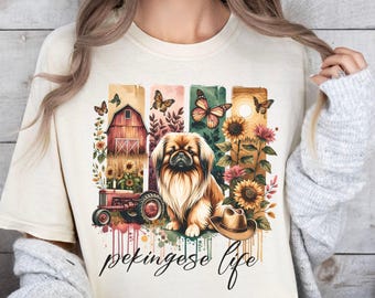 May include: A cream-colored t-shirt with a colorful graphic design featuring a Pekingese dog, a red tractor, sunflowers, and butterflies. The text "Pekingese Life" is printed below the graphic.