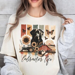 May include: Cream-colored t-shirt featuring a Labrador Retriever design with sunflowers, butterflies, a tractor, and a barn. The words "Labrador Life" are written in a script font below the graphic.