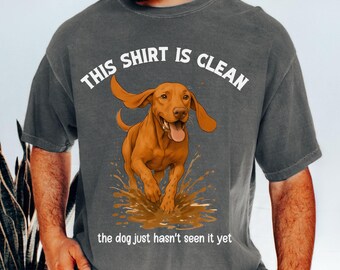 May include: Dark grey t-shirt featuring the text "THIS SHIRT IS CLEAN" above a graphic of a brown dog running through mud. The text "the dog just hasn't seen it yet" is below the dog graphic.