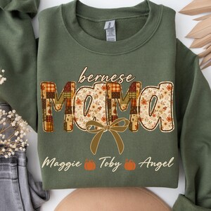 May include: An olive green crewneck sweatshirt featuring the text "bernese MAMA" in a large, patterned font with a plaid and floral design. A tan bow graphic sits below the text, followed by the names "Maggie", "Toby", and "Angel" with pumpkin icons.