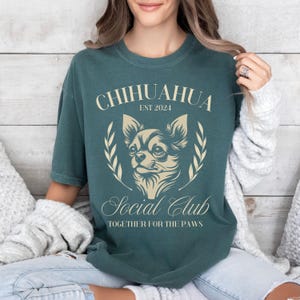 May include: Teal blue t-shirt with a graphic of a chihuahua in a wreath with the text "Chihuahua Est 2024 Social Club Together for the Paws".
