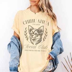 May include: A light yellow t-shirt with a graphic of a chihuahua dog. The graphic says "Chihuahua Social Club Est 2024 Together for the Paws".