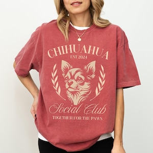 Woman standing with one hand on her leg and the other on her hip. She’s showing off her crimson red t-shirt. The design on her shirt is a Chihuahua dog “Social Club Together for the Paws” with personalized Est. year.