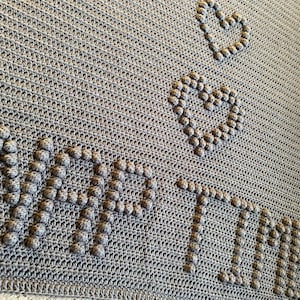 Può includere: Coperta in crochet grigia con le parole "nap time" e due cuori realizzati con punti in rilievo.