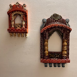 May include: Two decorative wall-mounted window frames featuring arched tops and intricate carvings. The smaller frame is orange with gold accents, while the larger frame is dark brown with gold and blue details. Both feature small hanging pegs at the base.