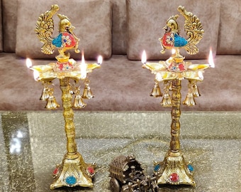 Pode incluir: Dois castiçais de metal dourado em forma de pavão com detalhes em vermelho, azul e verde. Cada castiçal tem cinco porta-velas e uma fila de sinos pendurados na base.