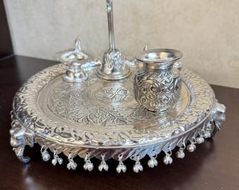 Puede incluir: Una bandeja de plata con intrincados diseños florales y un borde de cuentas. La bandeja contiene un diya de plata, una campana de plata y una copa de plata.
