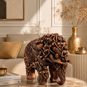 May include: A bronze-colored elephant sculpture with intricate carvings of figures. The elephant is standing on a marble-topped table. The sculpture is a decorative piece, likely for home decor.
