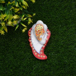 Könnte beinhalten: Ein rotes, weißes und goldenes Dekorationsobjekt in Form eines Blattes mit einer Darstellung eines Elefantenkopfes in der Mitte. Das Objekt ist mit Strasssteinen verziert.