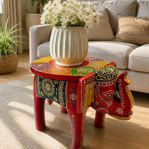 May include: A red wooden side table shaped like an elephant with a round top featuring colorful painted floral patterns. The table has four legs and a trunk extending from the front. A white ceramic vase filled with white daisies sits on top.