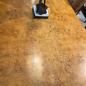 May include: A close-up of a wood table top with a burl wood veneer. The table top has a warm brown colour with a natural wood grain pattern. The table top is smooth and polished.