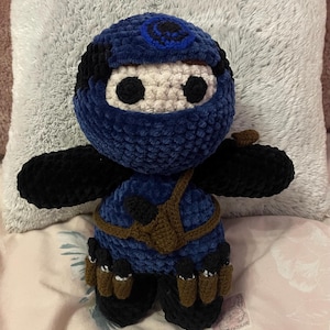 Puede incluir: Un muñeco de peluche tejido a ganchillo a mano con forma de ninja. La figura presenta una capucha azul y negra, una cara de color crema con ojos redondos negros, un torso azul, brazos negros y un cinturón marrón con pequeñas bolsas y accesorios.