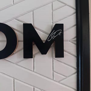 Puede incluir: Una obra de arte mural enmarcada con las letras "OM" en negro, sobre un patr&oacute;n geom&eacute;trico blanco. La letra "M" tiene una firma blanca. El marco es negro.