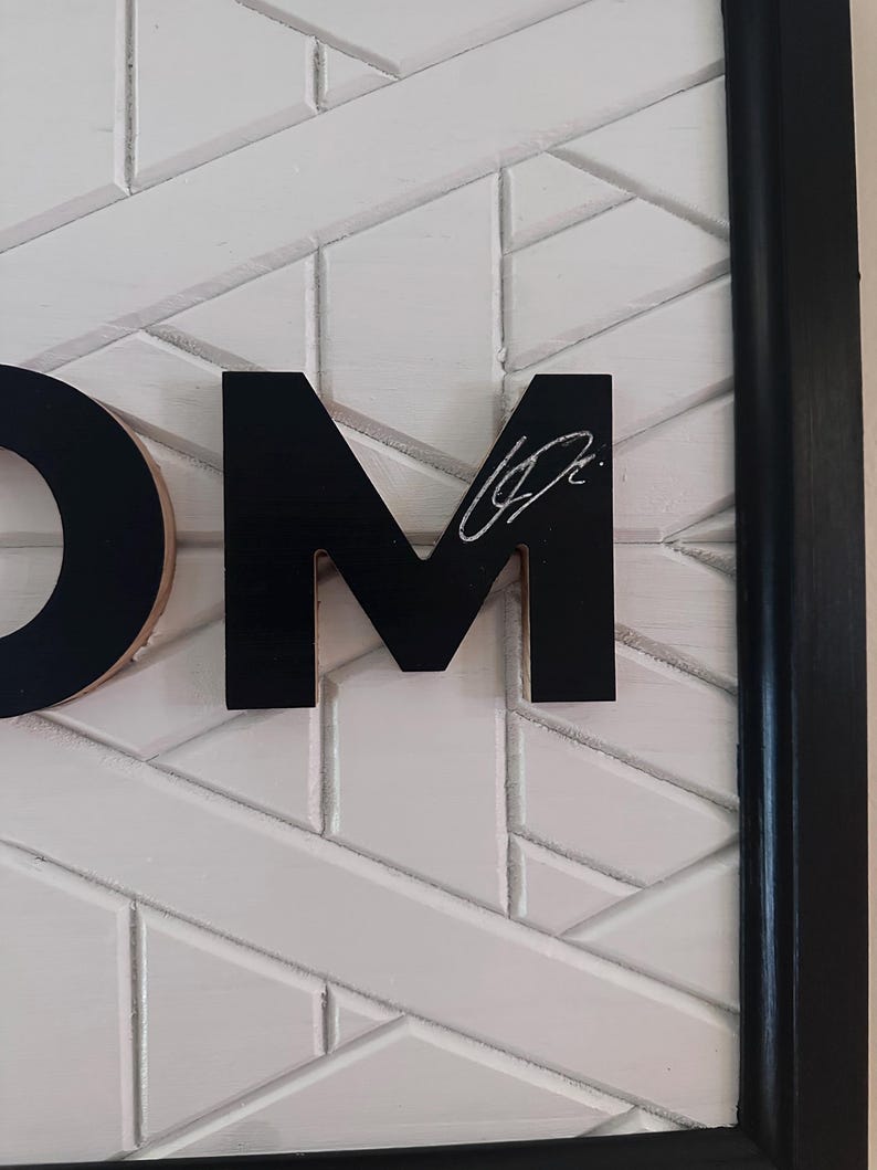 Puede incluir: Una obra de arte mural enmarcada con las letras "OM" en negro, sobre un patr&oacute;n geom&eacute;trico blanco. La letra "M" tiene una firma blanca. El marco es negro.
