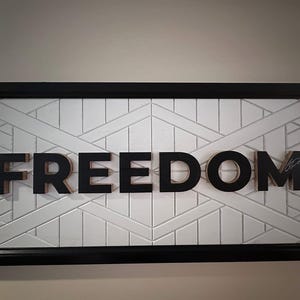 Puede incluir: Una obra de arte mural rectangular con la palabra "FREEDOM" en letras negras tridimensionales. Las letras est&aacute;n montadas sobre un fondo blanco con un patr&oacute;n geom&eacute;trico, enmarcado en negro.