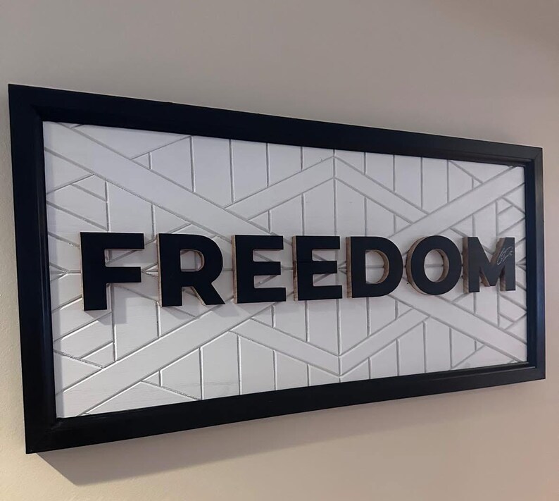 Puede incluir: Una obra de arte enmarcada con la palabra "FREEDOM" en letras negras y en negrita. Las letras est&aacute;n en relieve sobre un fondo blanco con un patr&oacute;n geom&eacute;trico. El marco es negro.