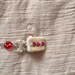 May include: A charm bracelet featuring a miniature cake pendant with strawberry accents. The bracelet also includes a red flower charm and a white bow. The background is a textured, light pink fabric.