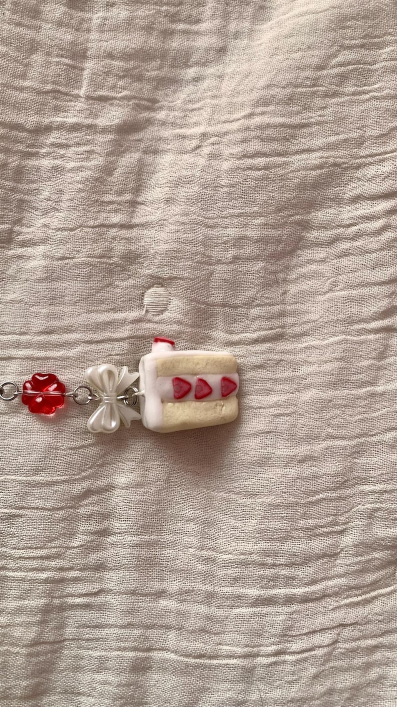 May include: A charm bracelet featuring a miniature cake pendant with strawberry accents. The bracelet also includes a red flower charm and a white bow. The background is a textured, light pink fabric.