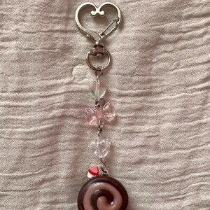 May include: A silver-toned keychain with a heart-shaped clasp. The keychain features clear heart-shaped beads, a pink bow-shaped bead, and a miniature chocolate roll charm. The charm has a swirl design and a small red and white heart.