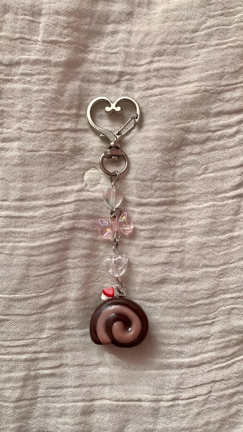 May include: A silver-toned keychain with a heart-shaped clasp. The keychain features clear heart-shaped beads, a pink bow-shaped bead, and a miniature chocolate roll charm. The charm has a swirl design and a small red and white heart.